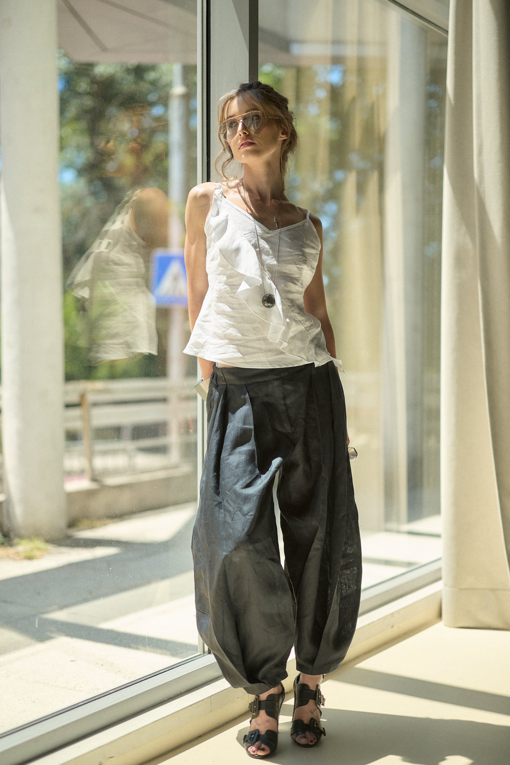 A woman wearing a white sleeveless top and dark baggy pants stands by a large window in a sunlit indoor setting.