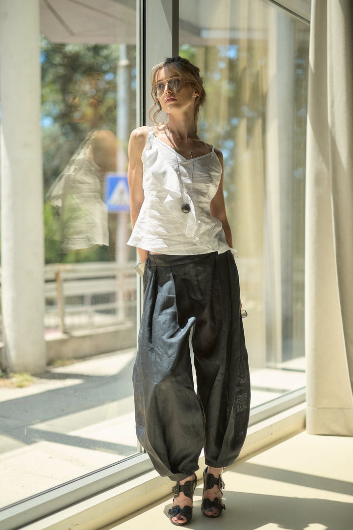 A woman wearing a white sleeveless top and dark baggy pants stands by a large window in a sunlit indoor setting.