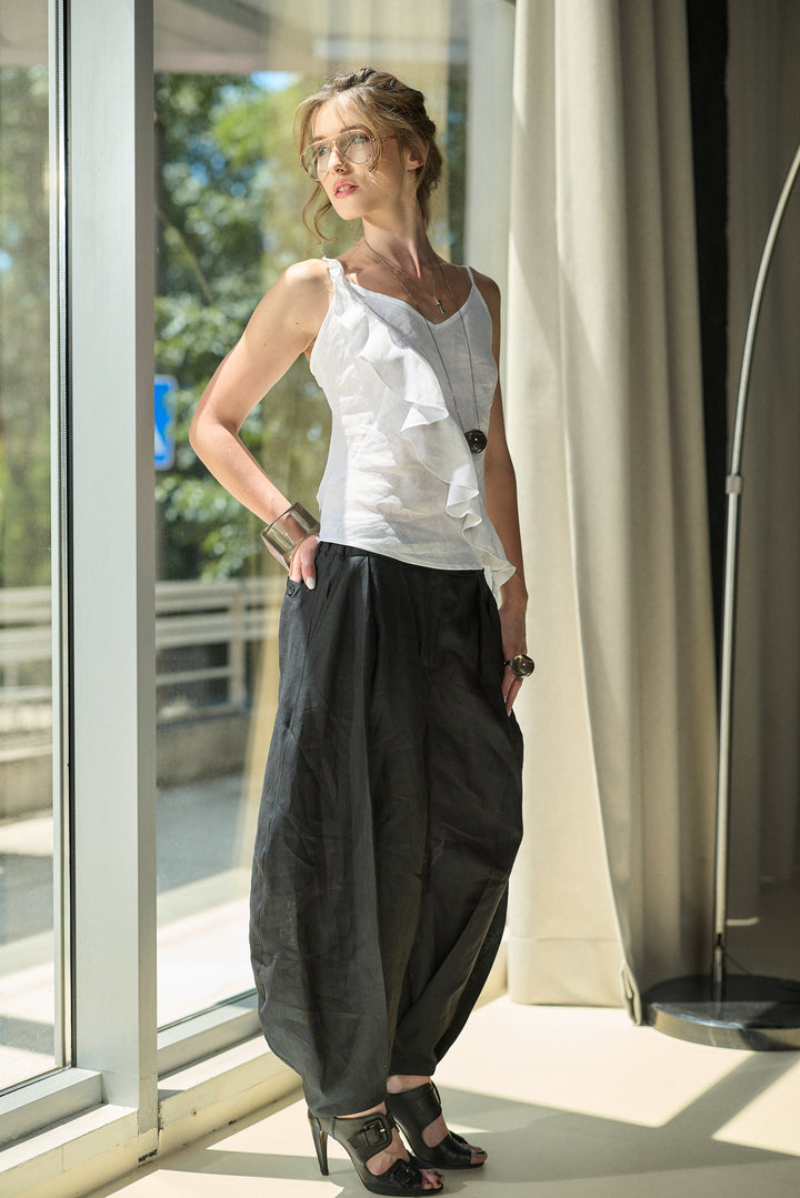 A woman wearing a white sleeveless ruffled top and loose black harem-style pants stands by a large window in a bright indoor setting.