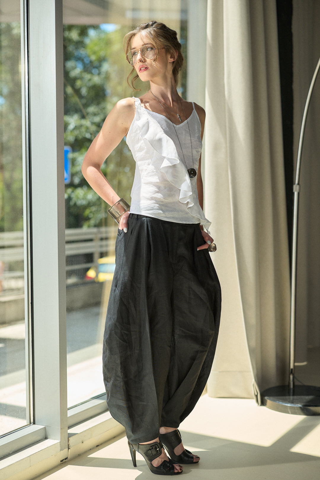 A woman stands by a large window wearing a white ruffled sleeveless top, loose black harem-style pants, and black heeled sandals in a bright indoor setting.