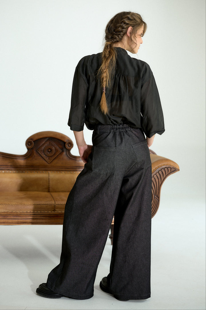 A woman stands with her back to the camera wearing a black three-quarter sleeve top and dark charcoal wide-leg trousers, posed in front of a light gray studio background with a wooden bench.