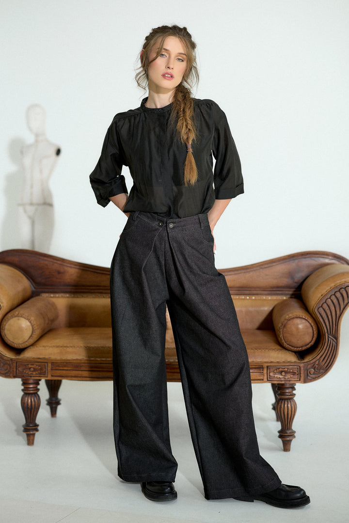 A woman stands in a studio wearing a black long-sleeve blouse and dark wide-leg trousers, posed in front of a brown wooden bench against a white background.