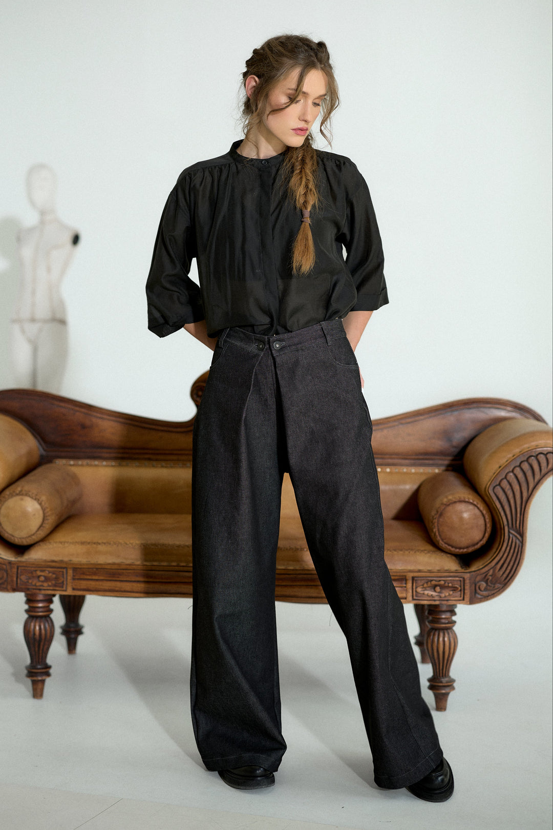 A woman stands in a studio wearing a black long-sleeve blouse and black wide-leg trousers, posed in front of a brown leather bench with a white mannequin in the background.