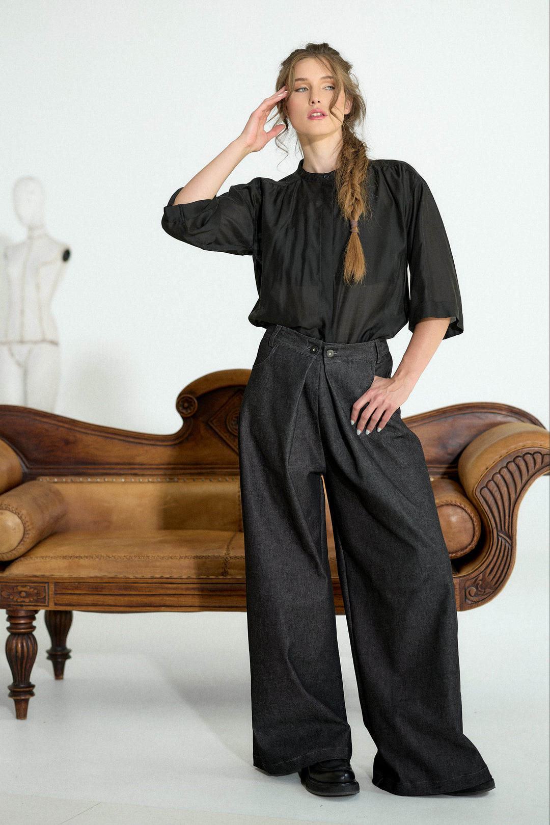 A model stands in a studio setting wearing a black short-sleeve blouse and wide-leg charcoal gray trousers, posed next to a brown leather chaise lounge.