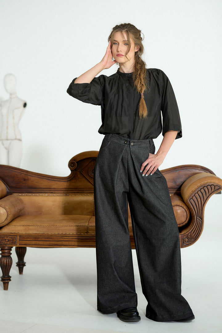 A woman stands in a studio wearing a black short-sleeve blouse and wide-leg charcoal gray trousers, posed next to a vintage wooden chaise lounge. A white mannequin is visible in the background against a white studio wall.