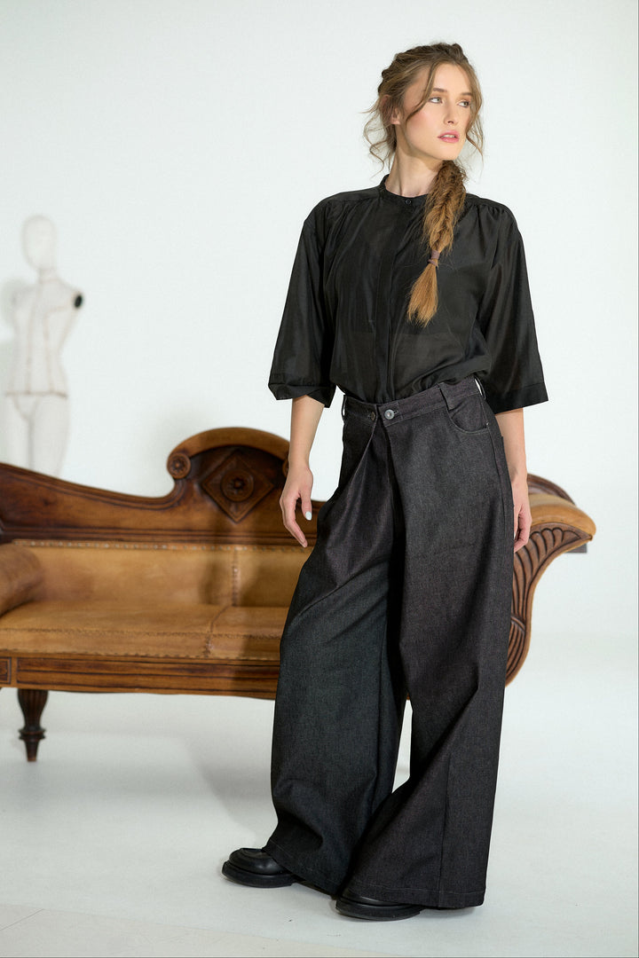A woman stands in a studio wearing a loose black short-sleeve top and very wide-leg dark gray trousers, posed in front of a wooden chaise lounge and a white mannequin on a white background.