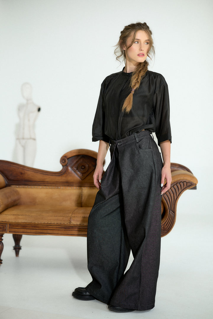 A woman stands in a studio wearing a black long-sleeve blouse and wide-leg dark gray trousers, posed in front of a wooden bench against a white background.