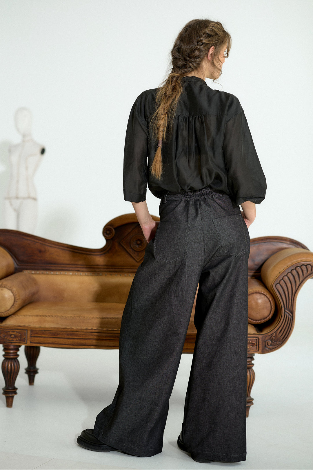 A woman stands with her back to the camera wearing a black long-sleeve top and wide-leg dark trousers, posed in front of a brown wooden chaise lounge against a white studio background.