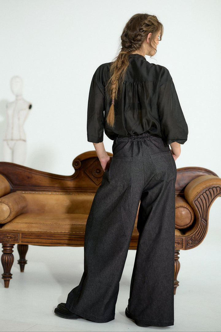 A woman stands with her back to the camera wearing a black long-sleeve top and wide-leg dark trousers, posed in front of a brown wooden chaise lounge against a white studio background.