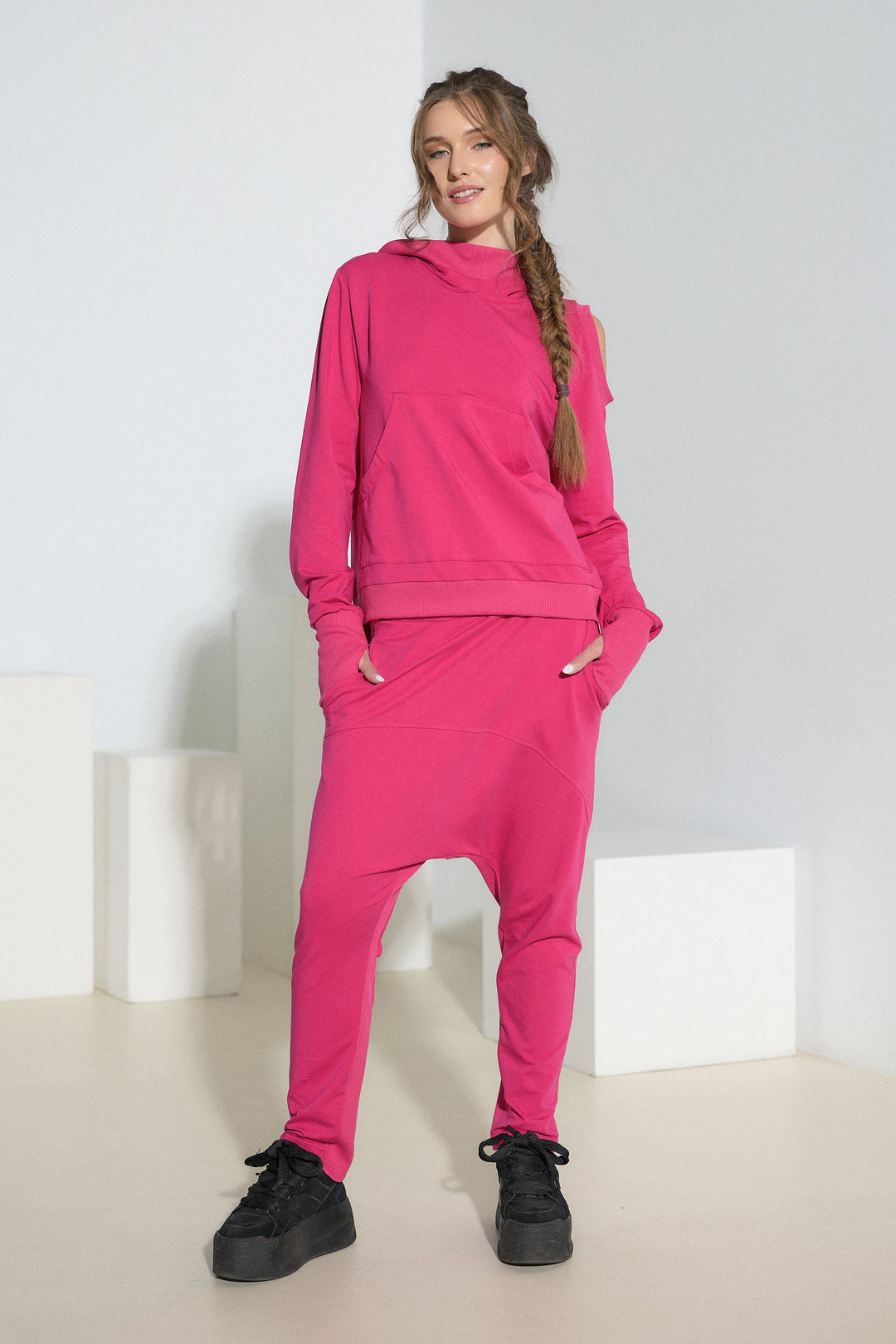A woman modeling a bright pink two-piece loungewear set consisting of a pullover hoodie and matching jogger pants, styled with black sneakers in a minimalist white studio setting.