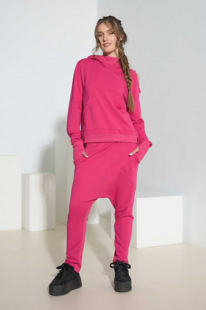 A woman modeling a bright pink two-piece loungewear set consisting of a pullover hoodie and matching jogger pants, styled with black sneakers in a minimalist white studio setting.