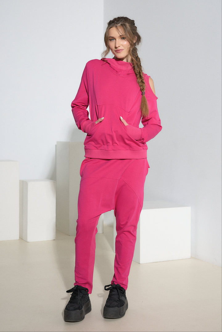 A woman modeling a bright pink matching lounge set consisting of a cold-shoulder pullover hoodie and jogger pants, paired with black platform sneakers, in a minimalist white studio setting.
