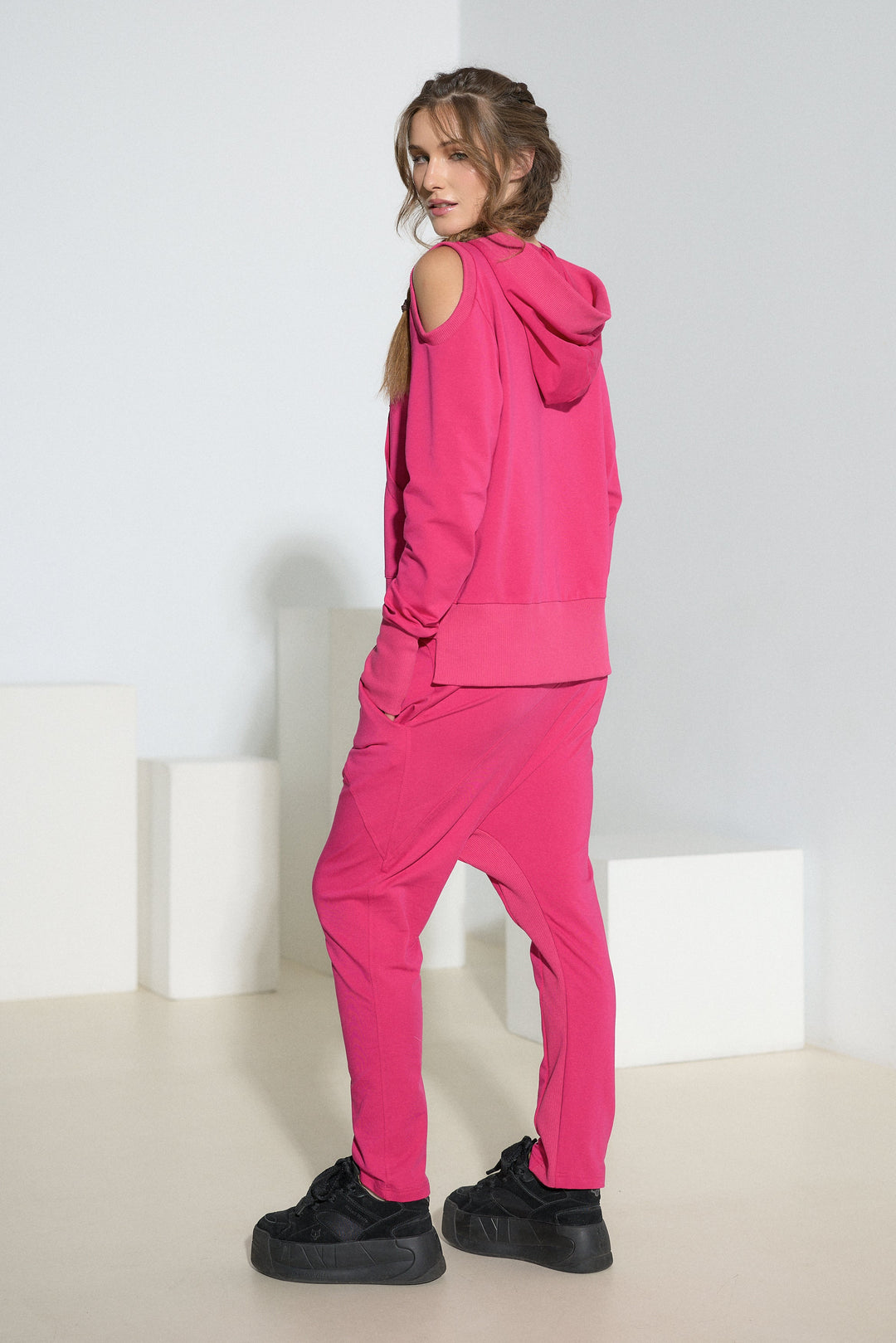 A woman modeling a bright pink matching lounge set consisting of a sleeveless hooded top and jogger pants, paired with black platform sneakers, standing in a minimalist white studio setting.