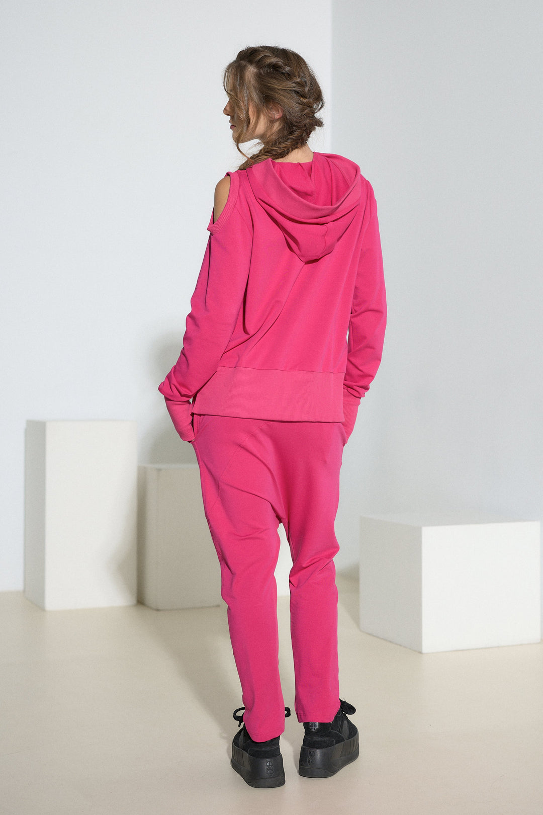 A model viewed from behind wearing a bright pink matching lounge set consisting of a hooded sweatshirt and jogger pants, styled with black platform sneakers in a minimalist studio setting.