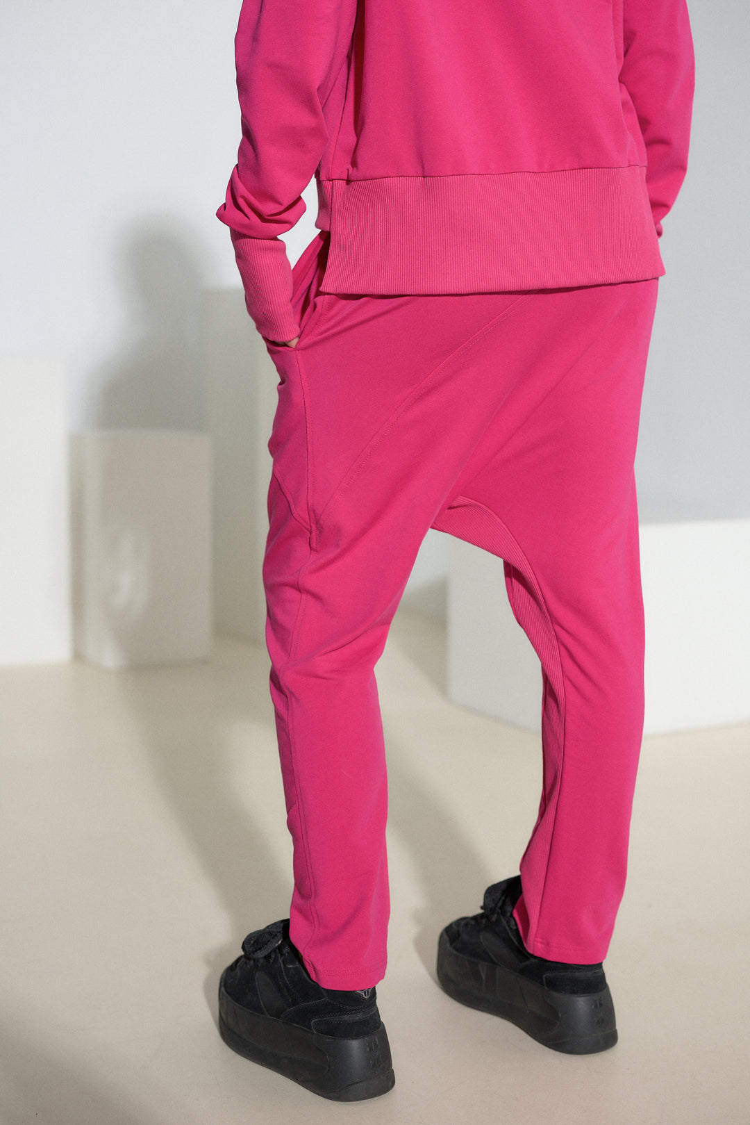 A person shown from the back wearing a bright pink matching tracksuit with baggy harem-style pants and black platform sneakers, standing in a minimalist studio setting.