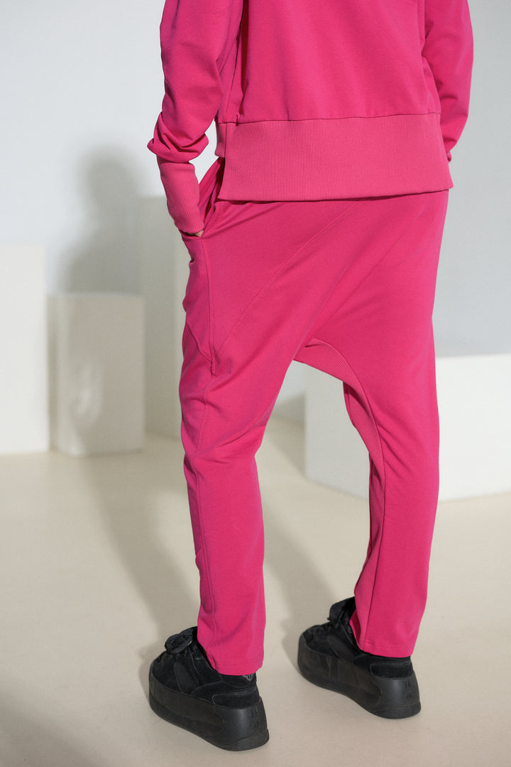 A person shown from the back wearing a bright pink matching tracksuit with baggy harem-style pants and black platform sneakers, standing in a minimalist studio setting.