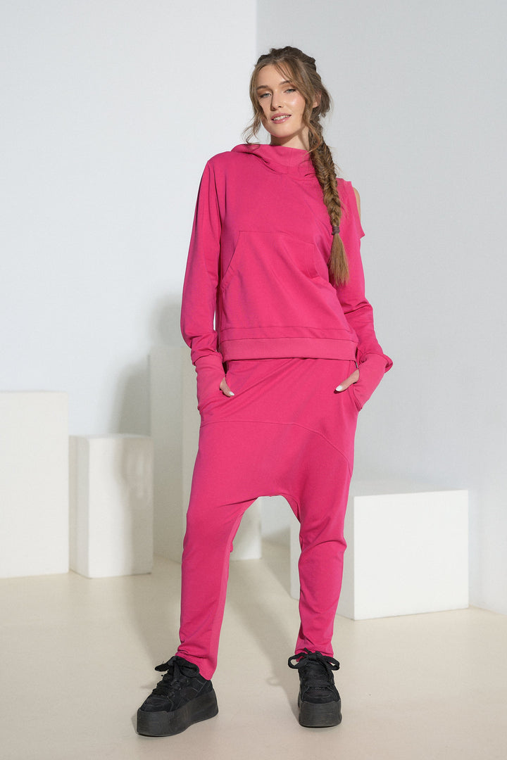 A woman modeling a bright pink two-piece loungewear set consisting of a pullover hoodie and matching jogger pants, styled with black sneakers in a minimalist white studio setting.