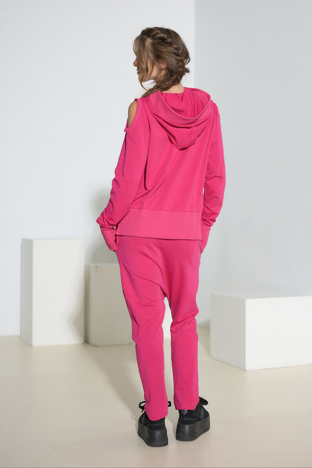 A model viewed from behind wearing a bright pink matching lounge set consisting of a hooded sweatshirt and jogger pants, styled with black platform sneakers in a minimalist studio setting.