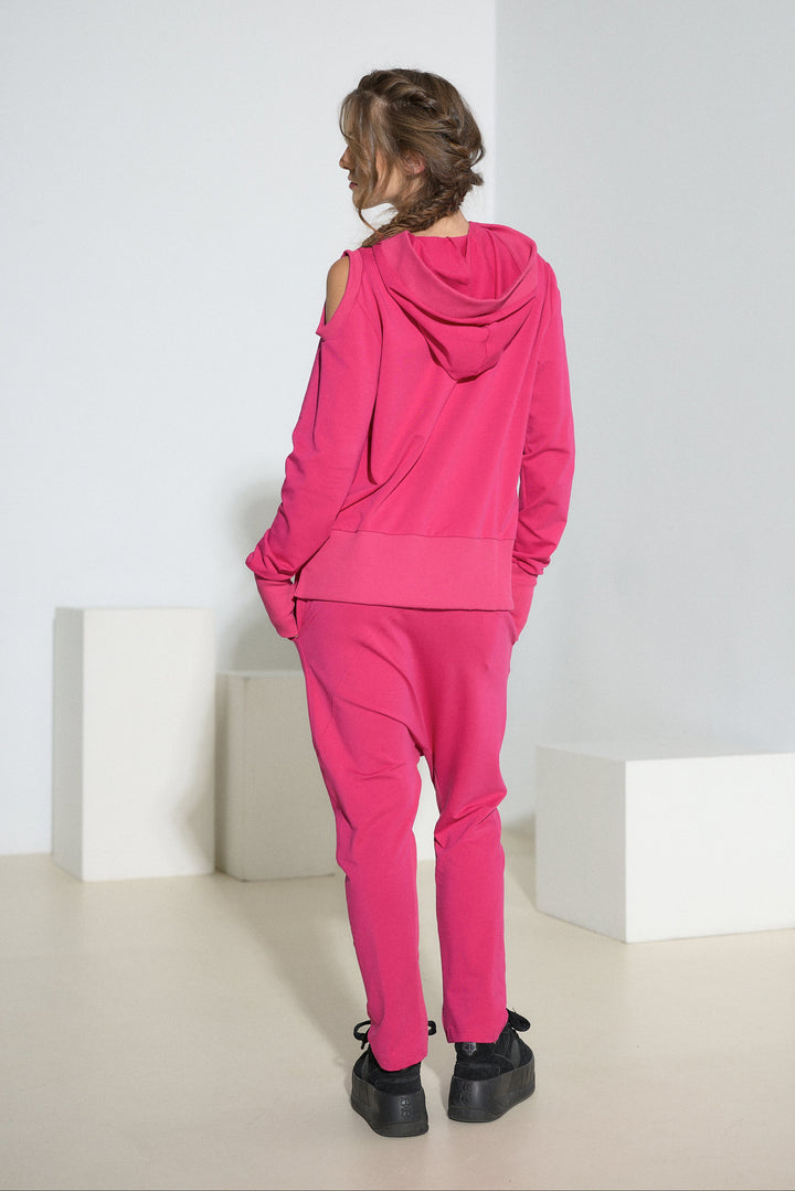 A model viewed from behind wearing a bright pink matching lounge set consisting of a hooded sweatshirt and jogger pants, styled with black platform sneakers in a minimalist studio setting.
