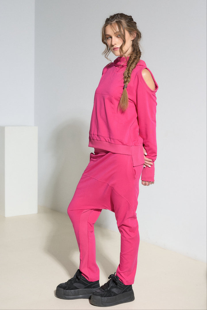 A model wearing a bright pink matching two-piece tracksuit with a cold-shoulder pullover hoodie and jogger pants, paired with black platform sneakers, in a minimalist studio setting.